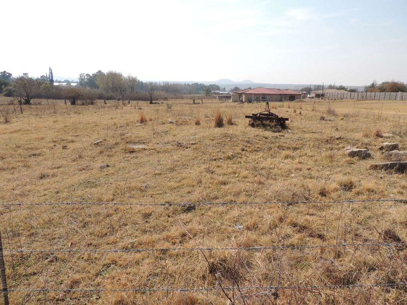 A view of a vacant residential plot featuring dry grass and perimeter barbed wire fencing with neighboring properties visible in the distance.