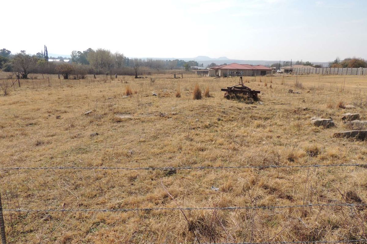 A view of a vacant residential plot featuring dry grass and perimeter barbed wire fencing with neighboring properties visible in the distance.