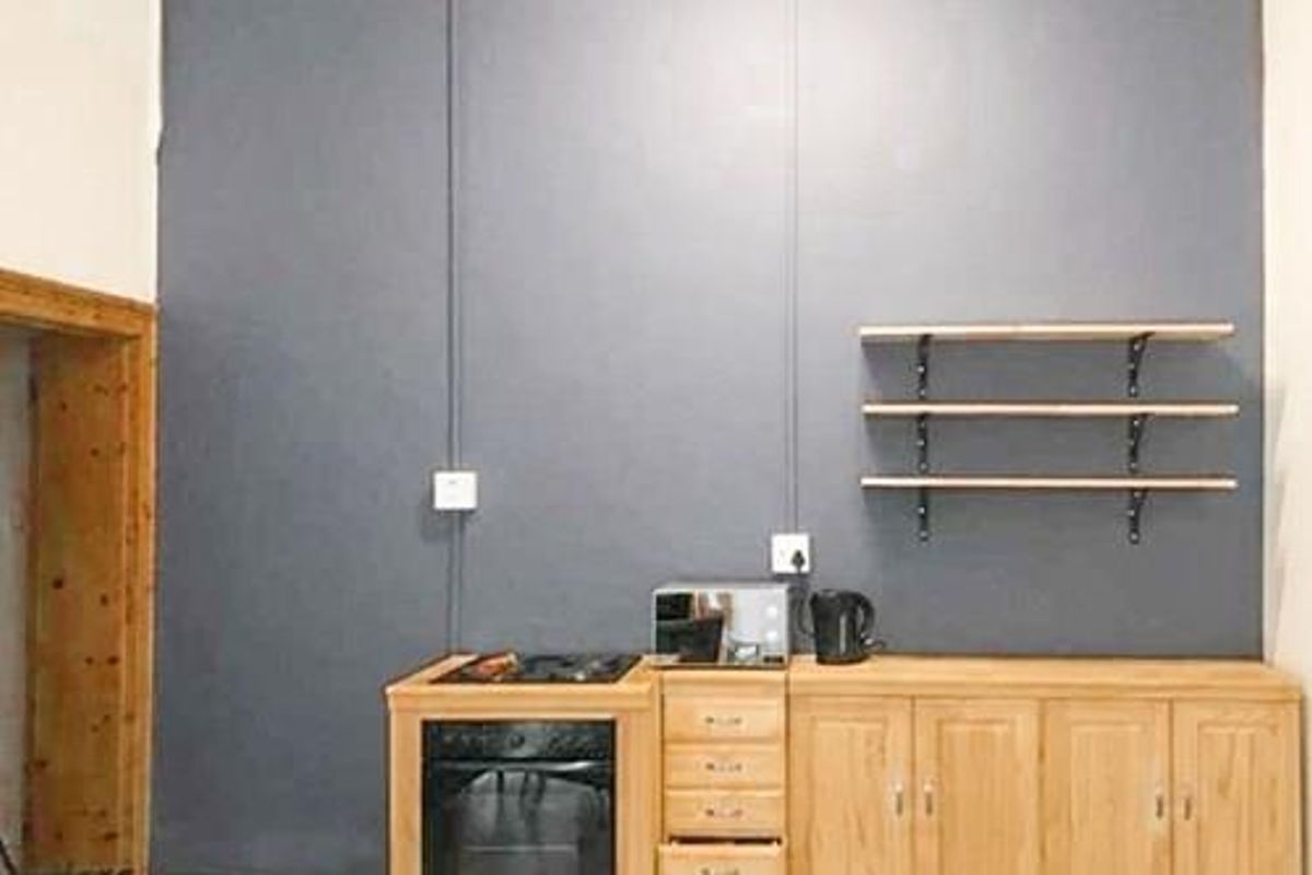 A compact kitchen area featuring wooden cabinetry, a built-in oven, a hob, and wall-mounted shelving against a dark grey feature wall.