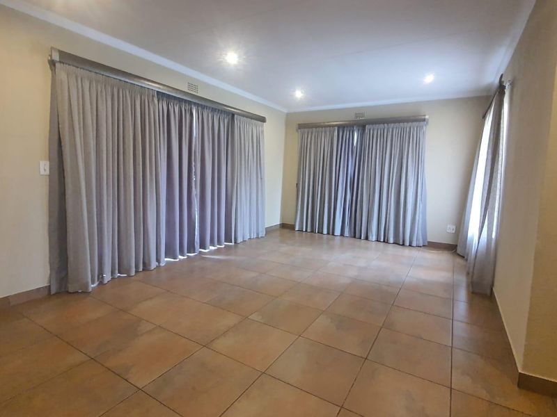 A spacious, empty living area featuring neutral tiled flooring, recessed downlighting, and large windows dressed with full-length curtains.