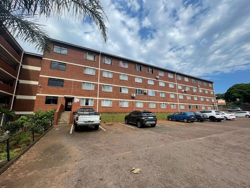Exterior view of a multi-story brick apartment building featuring a paved parking area and established landscaping.