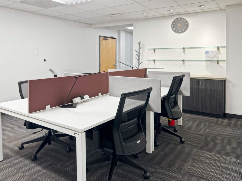 A professional office space featuring modular workstations, built-in storage cabinetry, glass shelving, and modern carpet tiles under a suspended ceiling.