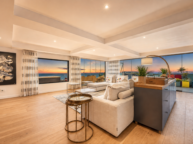 Spacious open-plan living area featuring high-end finishes, coffered ceilings, recessed lighting, and expansive glass doors opening to a balcony with panoramic sunset views.