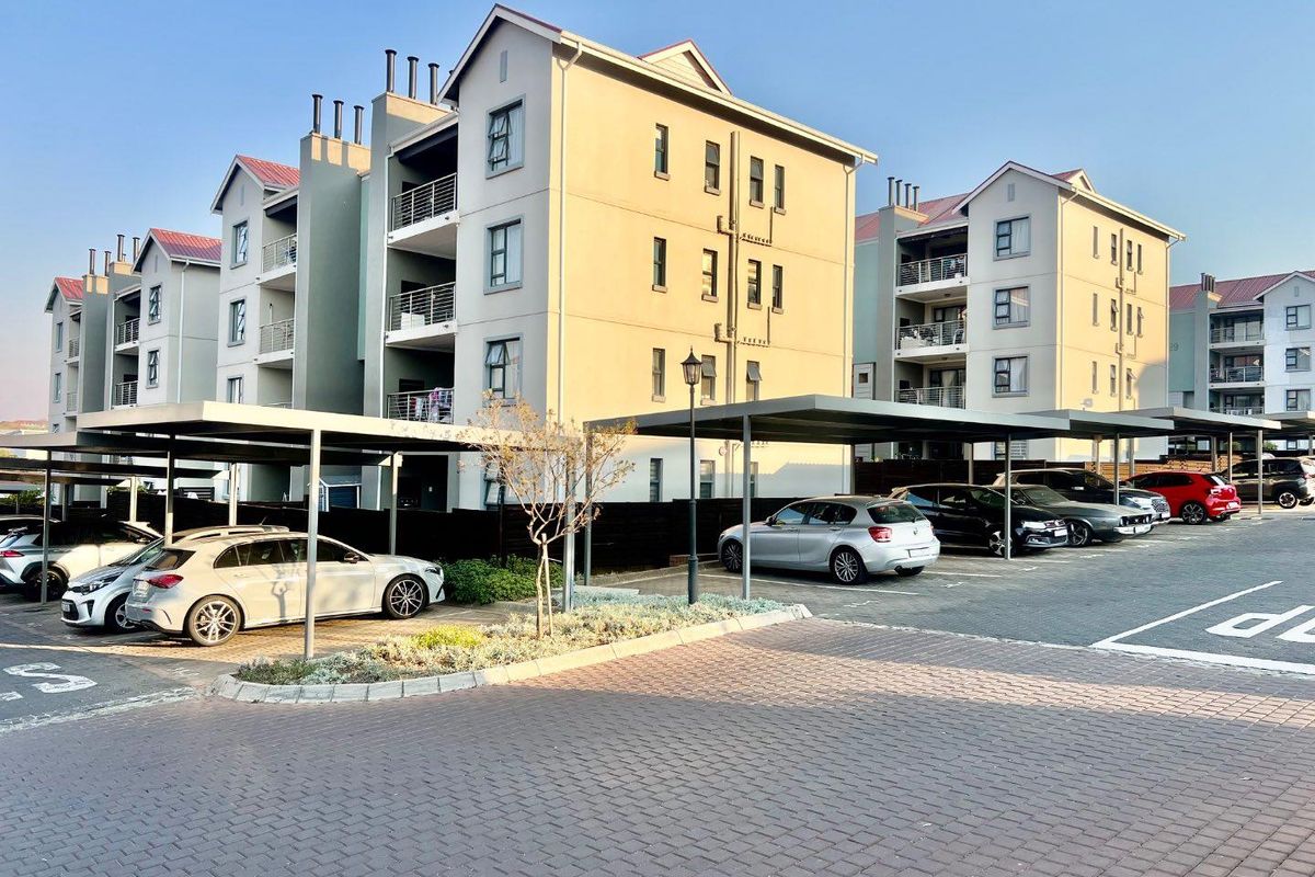 Exterior view of a modern apartment complex featuring covered parking bays, paved driveways, and multi-story residential units with private balconies.