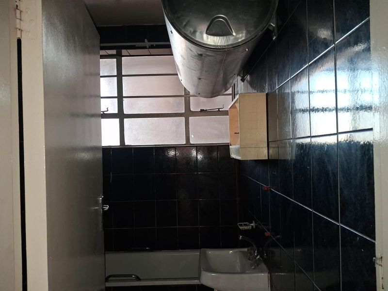 A dated bathroom featuring dark wall tiles, a standard bathtub, a wall-mounted basin, and an exposed electric water heater.