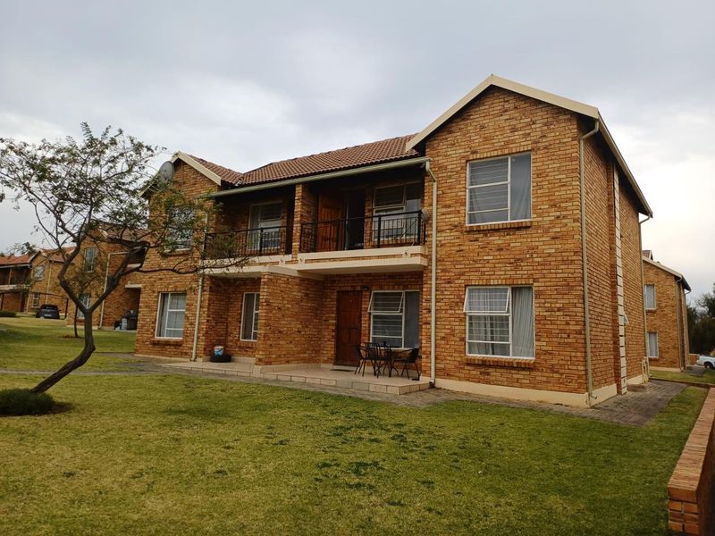Rear view of a two-story face brick apartment building featuring a ground-floor patio, upper-level balcony, and surrounding communal garden space.