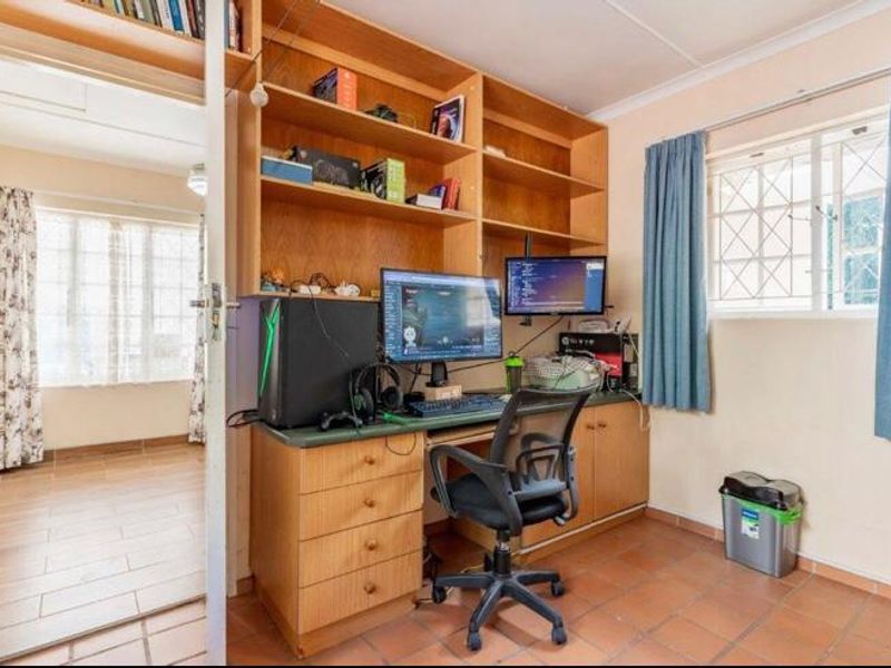 A functional home office featuring built-in wooden cabinetry, shelving, and tiled flooring, with a window providing natural light and security bars.