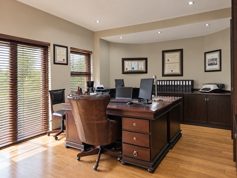 A well-appointed home office featuring warm timber flooring, built-in dark wood cabinetry, recessed lighting, and large windows with venetian blinds.