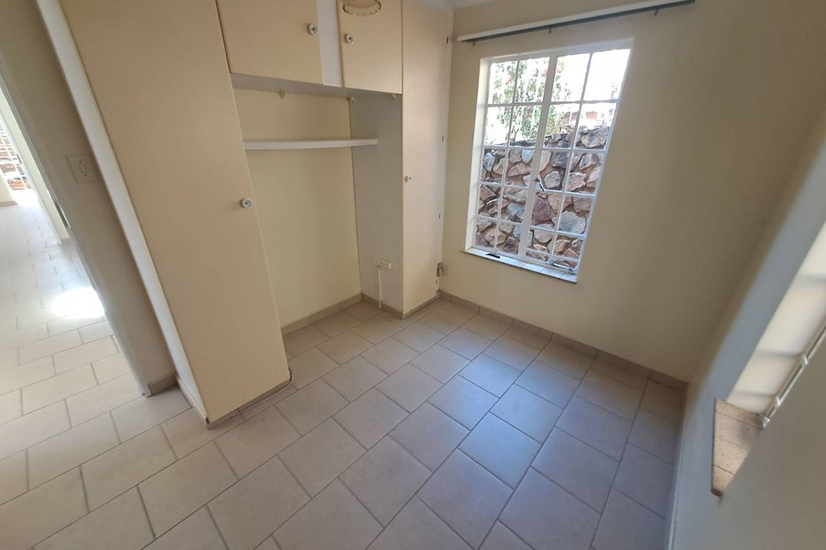 A compact, empty bedroom featuring tiled flooring, built-in cupboards, and a window with security bars overlooking a stone wall.