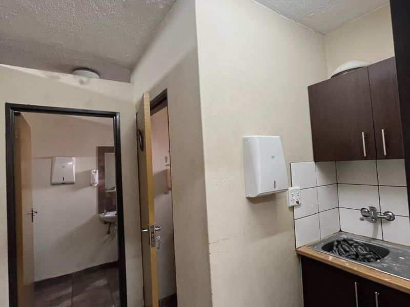 A compact kitchenette area featuring dark cabinetry, a stainless steel sink, and tiled backsplash, adjacent to a hallway leading to restroom facilities.
