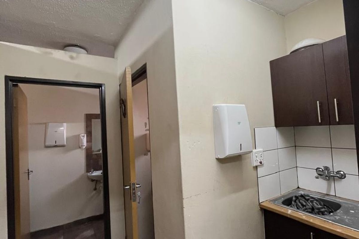 A compact kitchenette area featuring dark cabinetry, a stainless steel sink, and tiled backsplash, adjacent to a hallway leading to restroom facilities.