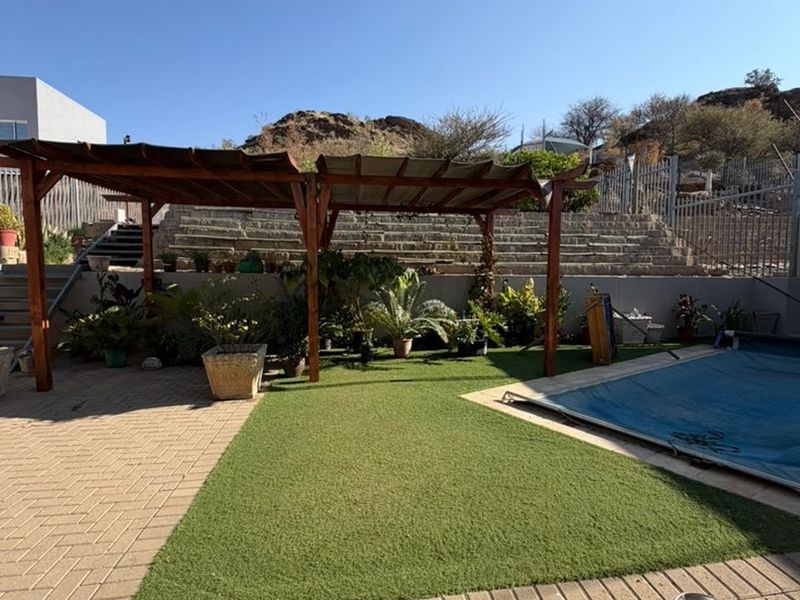 Outdoor pool area featuring a paved patio, artificial lawn, wooden pergola, and tiered stone retaining walls leading to the upper garden level.