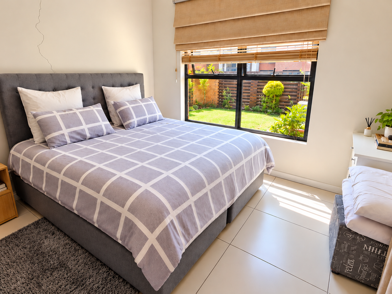 A bright bedroom featuring tiled flooring, a large window with roman blinds, and a view of the garden.