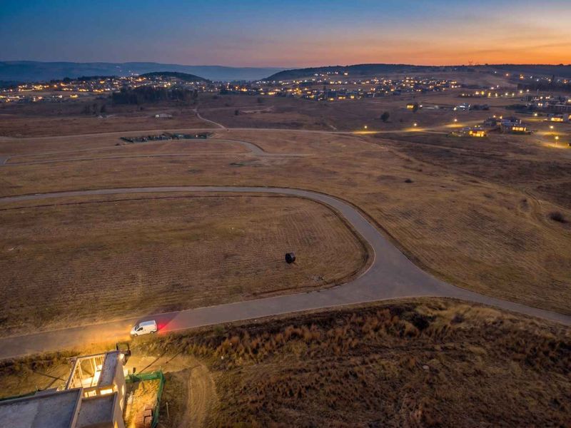 An aerial view of a residential development site at dusk, showcasing the surrounding landscape, road infrastructure, and nearby neighborhood lights.