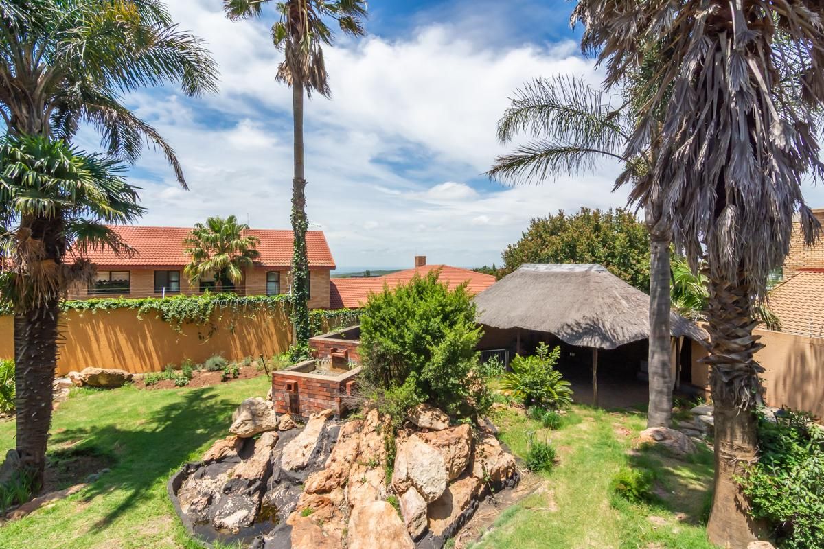 A spacious rear garden featuring a rockery water feature, established palm trees, and a traditional thatched lapa area for outdoor entertaining.