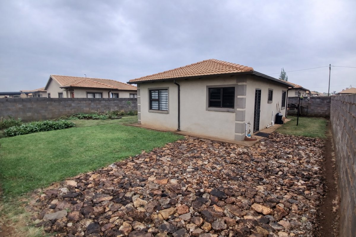 Rear view of the property featuring a tiled roof, plastered exterior walls, and a low-maintenance garden with a mix of lawn and stone landscaping.