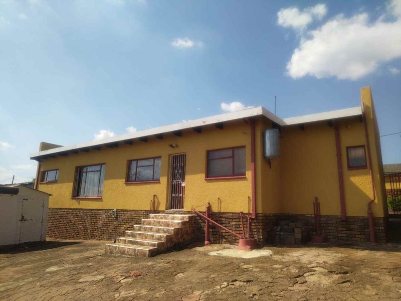 Rear view of a single-story house featuring a tiled staircase, brick plinth, and exterior geyser, set against a large, unpaved yard area.