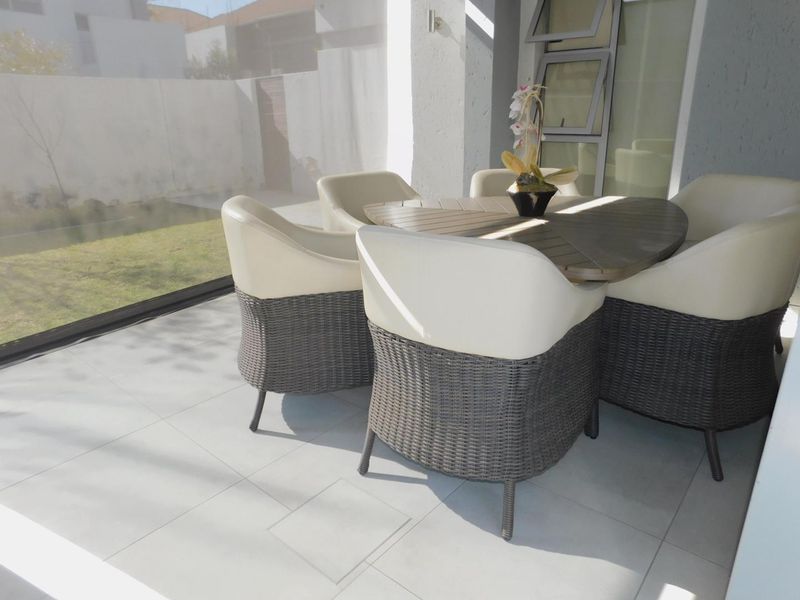 A covered patio area featuring light-toned floor tiles, aluminium window frames, and an outdoor roller blind, providing a seamless transition to the garden.