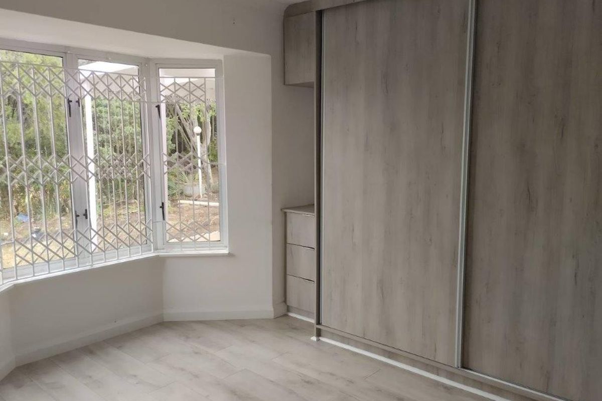 A bright, empty bedroom featuring modern laminate flooring, a large bay window with security gates, and a spacious built-in wardrobe with sliding doors.