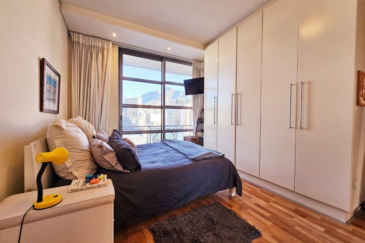 A well-lit bedroom featuring extensive built-in cupboards, warm laminate flooring, and a large window offering urban views.