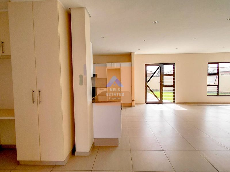 Spacious open-plan living and kitchen area featuring neutral tiled flooring, built-in cabinetry, and large windows providing ample natural light.