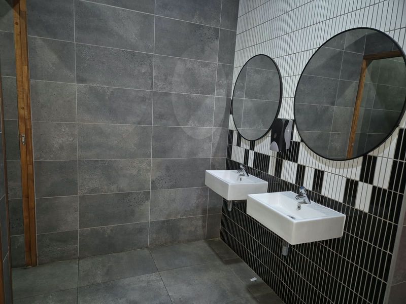 Modern commercial bathroom featuring dual wall-mounted ceramic basins, contemporary chrome fixtures, round mirrors, and stylish contrasting grey and black-and-white tiled wall finishes.
