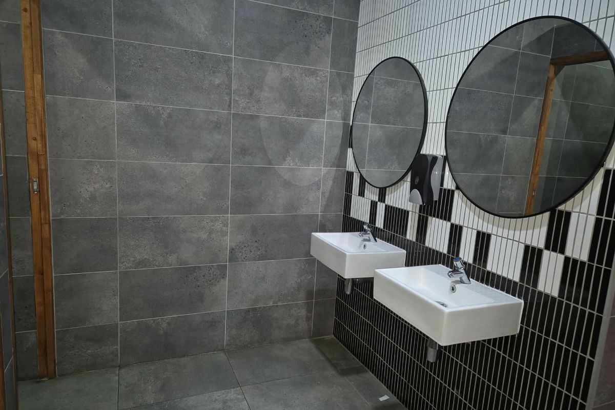 Modern commercial bathroom featuring dual wall-mounted ceramic basins, contemporary chrome fixtures, round mirrors, and stylish contrasting grey and black-and-white tiled wall finishes.
