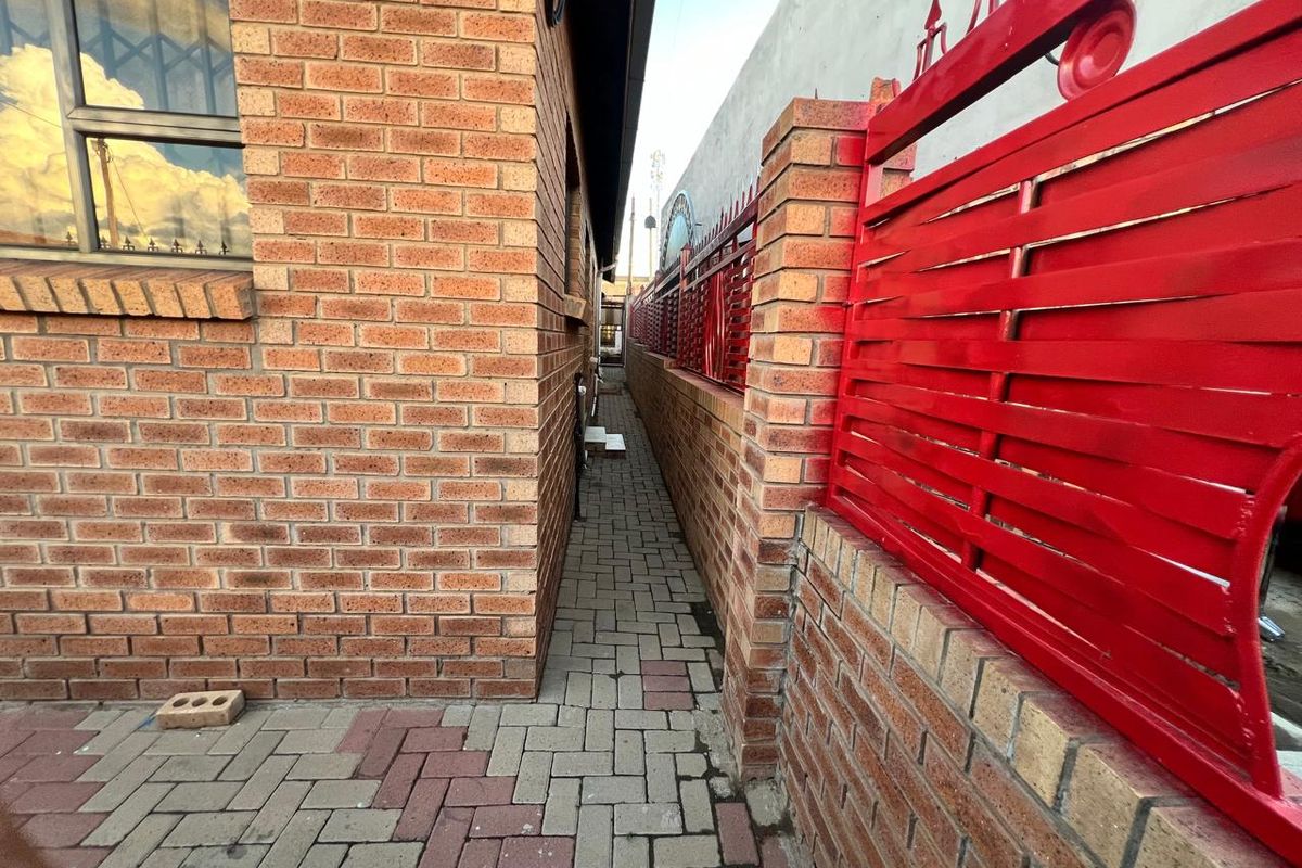 A narrow side passage featuring brick paving, a brick boundary wall, and red metal security fencing along the side of the house.