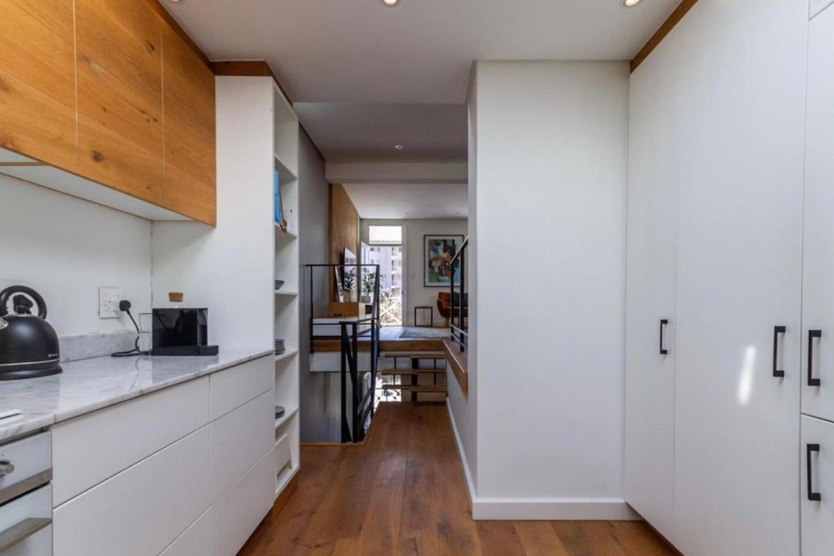 Modern kitchen featuring sleek white cabinetry, wood accents, stone countertops, and integrated appliances, leading into an open-plan living area with a staircase.