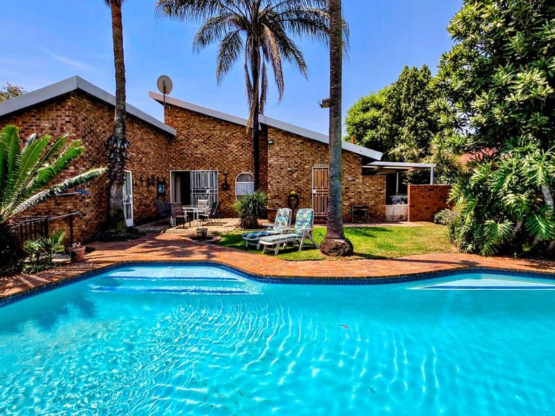A well-maintained backyard featuring a swimming pool, paved patio area, and established garden set against a classic brick house facade.