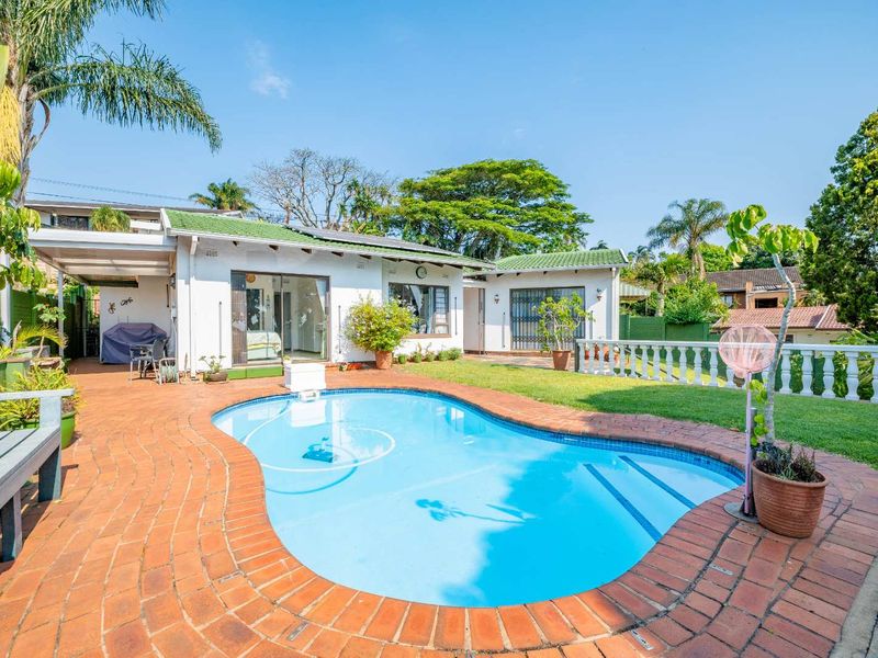 A well-maintained backyard featuring a swimming pool, brick-paved surround, covered patio area, and a landscaped lawn with a decorative balustrade.