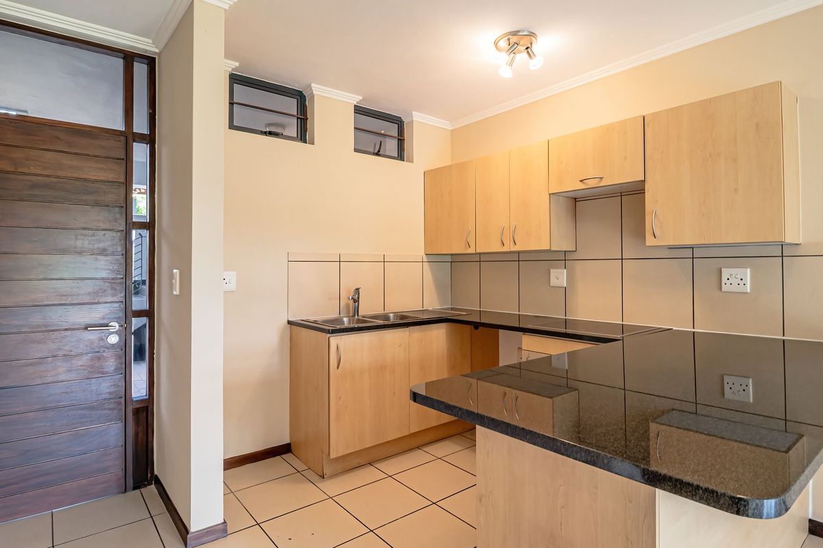 A clean, functional kitchen featuring light wood cabinetry, dark granite countertops, tiled backsplash, and ceramic flooring, with a solid timber entrance door.