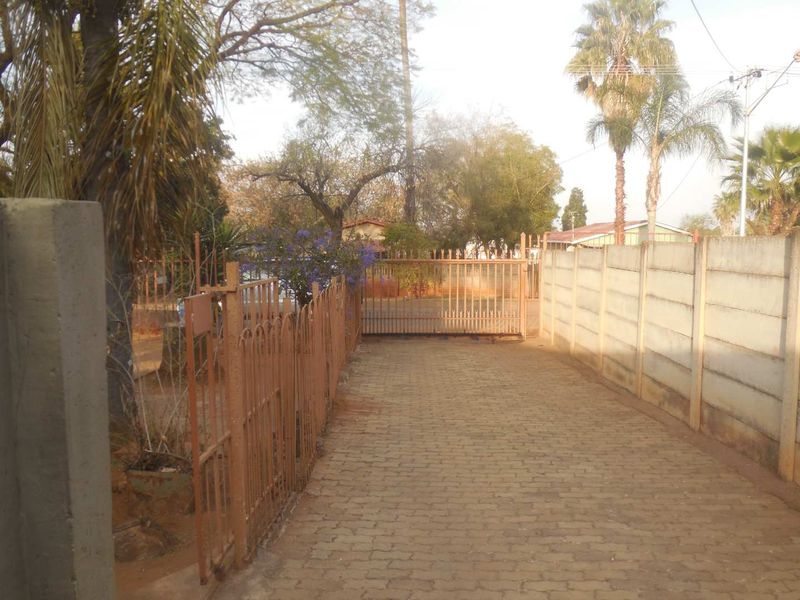 A paved driveway leading to a metal sliding gate, enclosed by a precast concrete boundary wall and metal fencing.