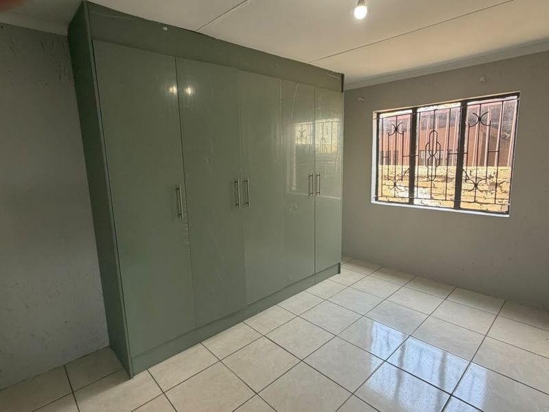 A bright, empty bedroom featuring tiled flooring, a large built-in wardrobe with sage green doors, and a window fitted with security bars.