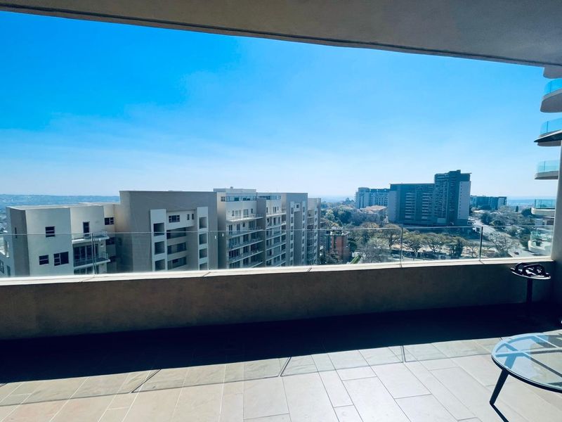 Spacious covered balcony featuring a modern glass balustrade and tiled flooring, offering expansive urban views.