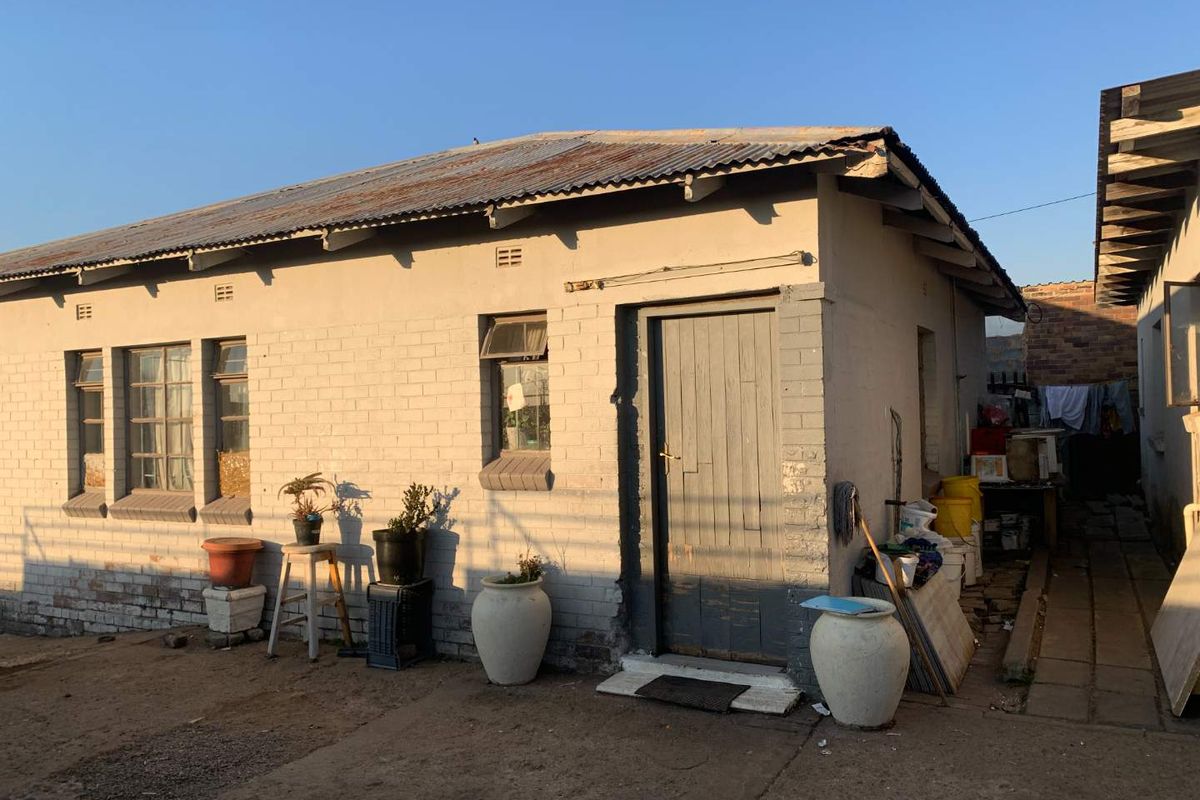 Exterior view of a single-story residential dwelling featuring a corrugated iron roof, brick masonry walls, and a side entrance door.