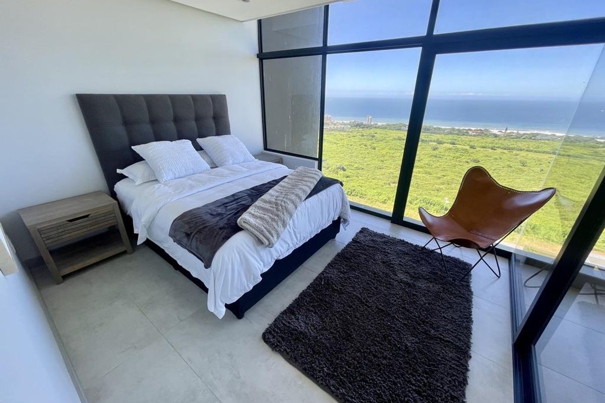 A modern bedroom featuring floor-to-ceiling windows with expansive coastal and landscape views, tiled flooring, and recessed ceiling lighting.