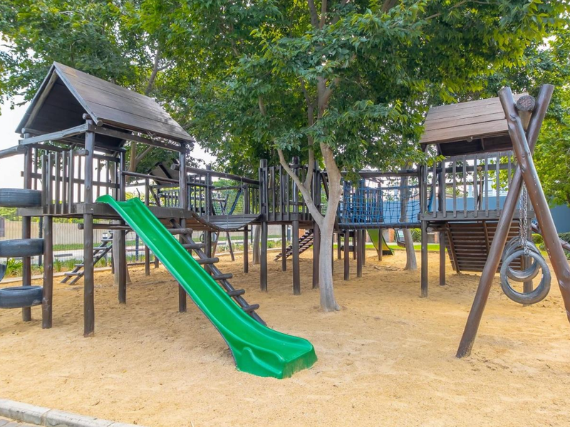 A communal outdoor playground area featuring a wooden climbing structure, slide, and swings set within a sand-filled play zone.