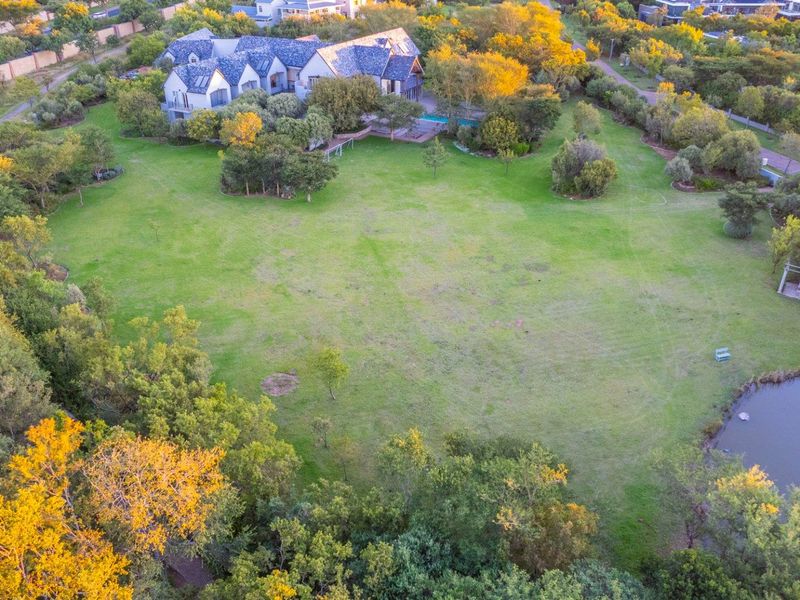 An aerial view of a large, well-maintained residential plot featuring expansive green lawns, mature trees, and a natural pond.