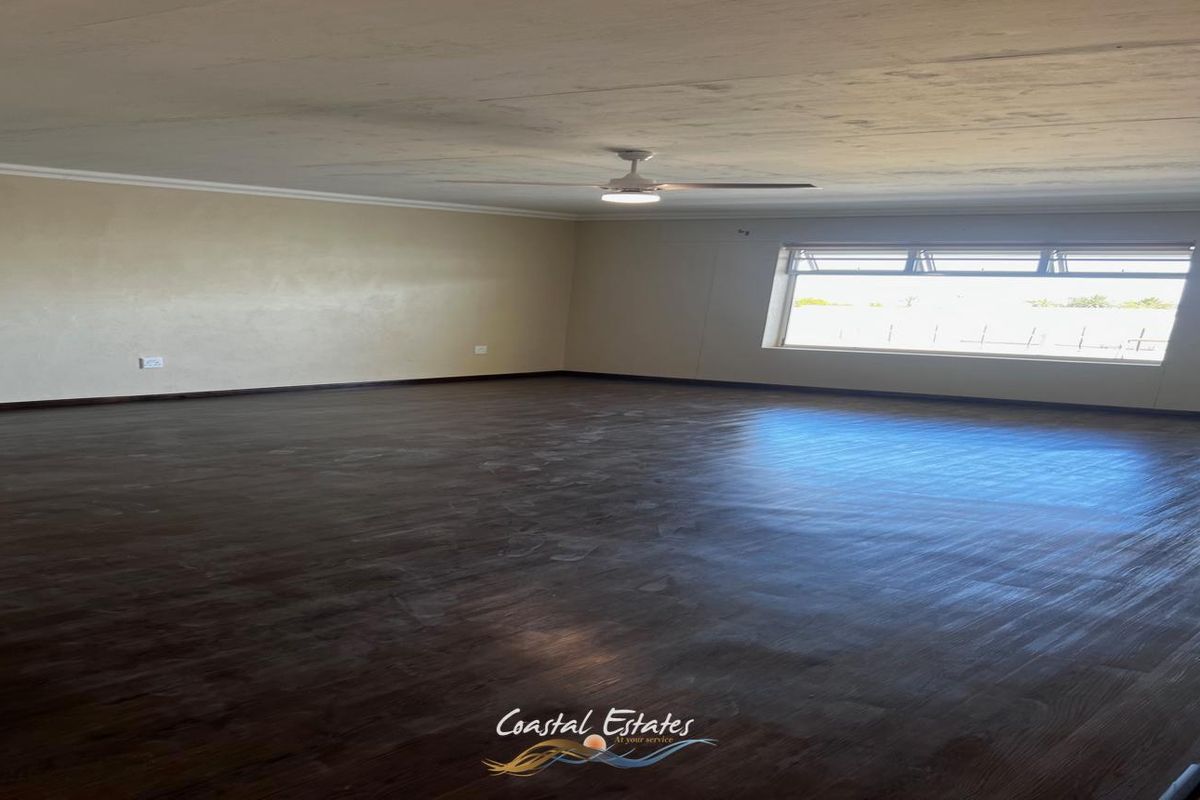 A spacious, empty living room featuring dark wood-look flooring, a ceiling fan, and large windows providing natural light.