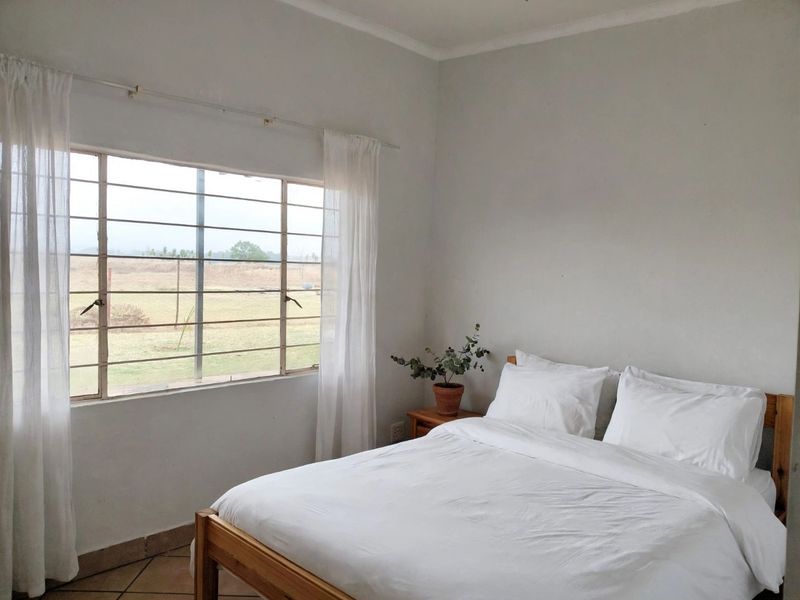 A simple bedroom featuring tiled flooring and a large window with security bars, offering a view of the surrounding landscape.