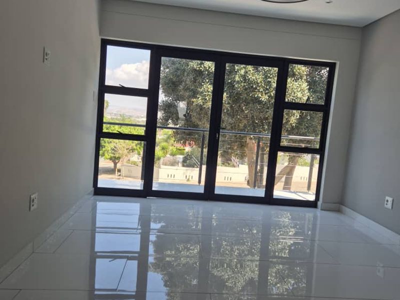 A bright, empty living room featuring polished large-format floor tiles, modern lighting, and large black-framed sliding doors leading to an outdoor area.