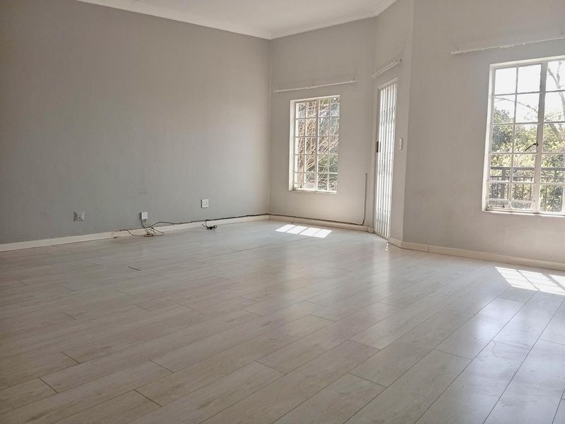 A bright, empty living room featuring light-toned laminate flooring, neutral walls, and large mullioned windows with an integrated security gate.