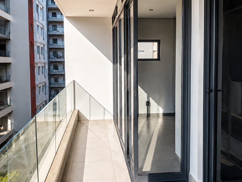 A modern, covered balcony featuring tiled flooring, a sleek glass balustrade, and large aluminium-framed sliding doors providing seamless indoor-outdoor flow.