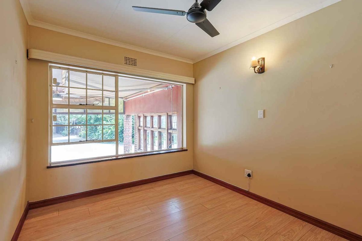 A bright, empty bedroom featuring light wood-look flooring, a ceiling fan, and a large window overlooking an outdoor patio area.
