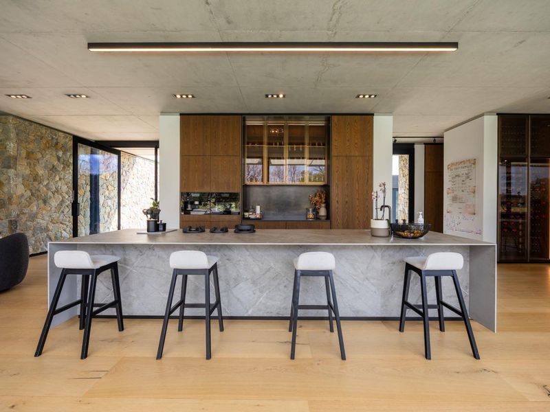 Modern open-plan kitchen featuring a large stone island, high-end cabinetry, integrated wine storage, and a striking exposed concrete ceiling with premium finishes.