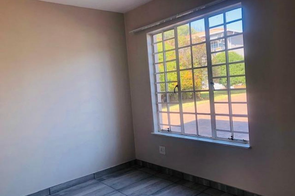 A bright, empty bedroom featuring modern grey wood-look floor tiles and a large window with security bars, offering a clean and neutral living space.