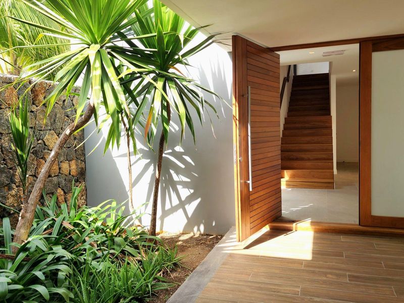 Modern entrance featuring a large timber pivot door, tiled patio area, and a stone-clad feature wall with landscaped garden.