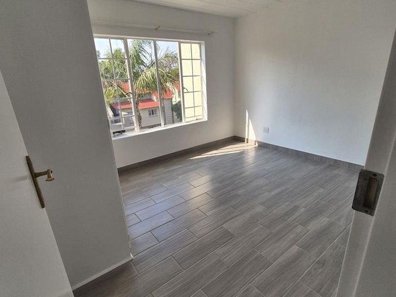 A bright, empty bedroom featuring modern wood-look floor tiles, a large window providing natural light, and a clean, neutral interior finish.