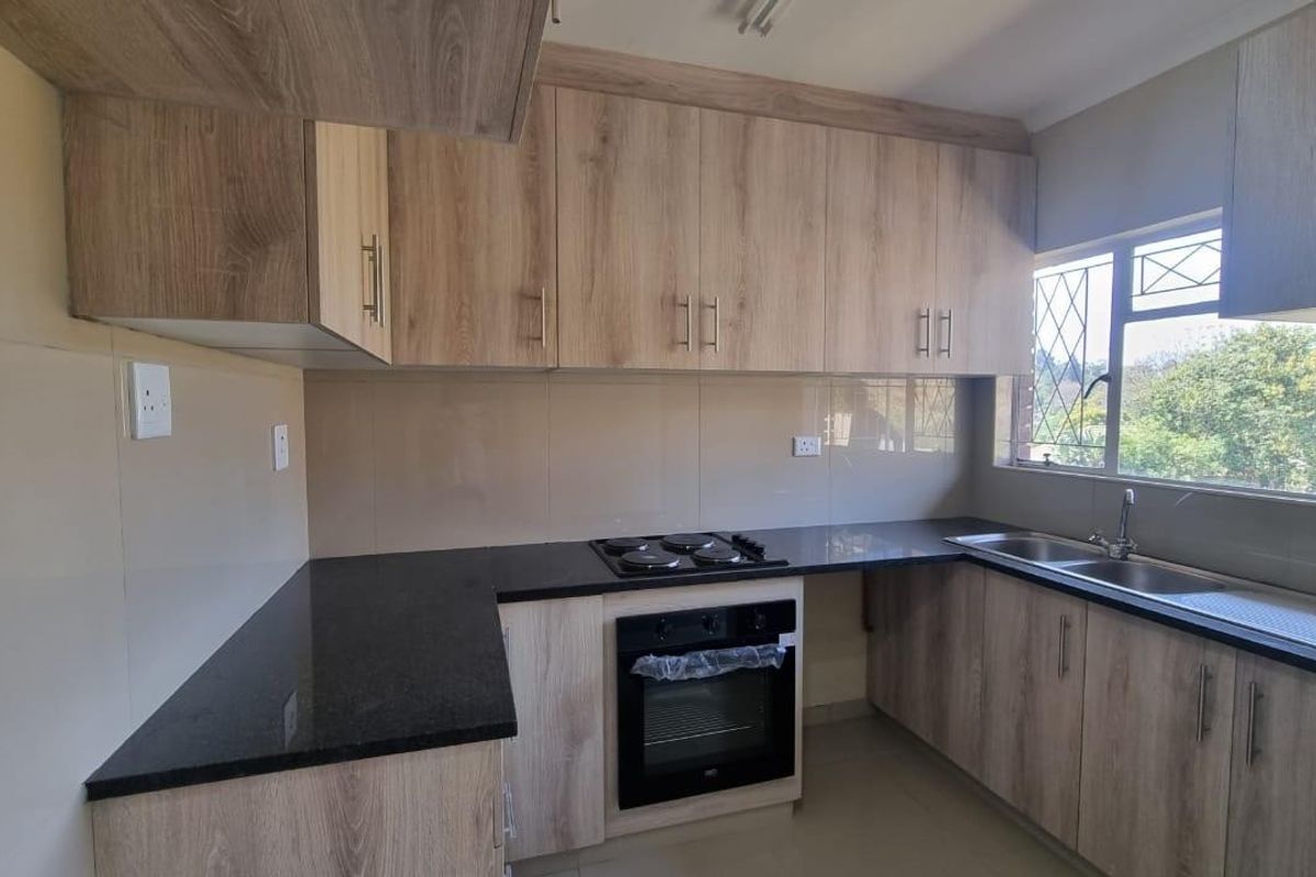A clean, modern kitchen featuring light wood-finish cabinetry, dark granite countertops, a built-in oven, electric hob, and a double sink with a window view.